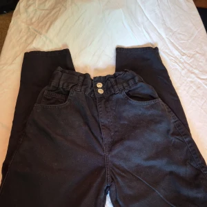Svarta byxor från Pull&Bear - Säljer ett par svarta byxor från Pull&Bear i storlek EUR 32 (USA 0, MEX 22). Byxorna har en högmidjad passform med elastisk midja och två knappar framtill. De har både fram- och bakfickor och är gjorda av ett bekvämt material som ser ut som jeans. Perfekta för både vardag och fest!