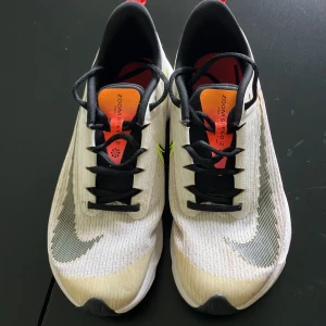 Nike ZoomX Sneakers - Säljer ett par grymma Nike ZoomX sneakers i vitt med svarta och orange detaljer. De har en snygg och modern design med snörning och en bekväm passform. Perfekta för träning eller vardagsbruk!