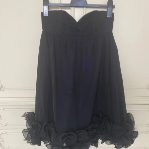 Svart axelbandslös klänning med volang  - Jättefin svart axelbandslös klänning med fyllig volang nertill i strlk 36 från H&M. Går tio mitten av låren (jag är 167 cm) och ger väldigt fint ”svall” då volangen håller formen rund nedtill. 