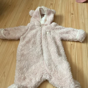 Rosa teddyoverall från Ellos - Säljer en supergullig rosa teddyoverall från Ellos. Den är perfekt för att hålla din bebis varm och mysig under kalla dagar. Overallen har en dragkedja framtill och en söt huva med små öron. Den är långärmad och har små tassavtryck på fötterna. Perfekt för höst och vinter!