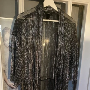 Glittrig långärmad klänning - Säljer en superglittrig långärmad kimono i silver. Perfekt för fest eller nyår! Den har en öppen design och är gjord i ett lätt, transparent material som ger en snygg effekt. Den är i storlek M och är i nyskick.