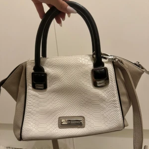 Vit handväska från Steve Madden - Säljer en superfin vit och beige handväska från Steve Madden. Den har ett snyggt ormskinnsmönster och svarta handtag. Väskan har en dragkedja upptill och insidan är randig i beige och vitt. Perfekt för både vardag och fest 💃