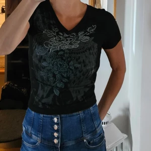 Svart t-shirt med blommönster -  Nyskick Säljer en svart t-shirt med ett snyggt blommönster och glittriga detaljer på framsidan. Den har en v-ringad hals och korta ärmar, perfekt för både vardag och fest. Passformen är tight och materialet känns mjukt och bekvämt.