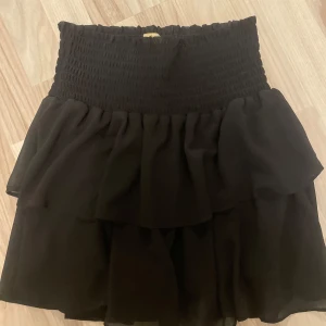 Svart kjol från Chelsea - Säljer en superfin svart kjol från Chelsea. Den har en smockad midja som ger en bekväm och flexibel passform. Kjolen är kort och har flera lager med volanger som ger en härlig volym och rörelse. Perfekt för både vardag och fest!😇😇