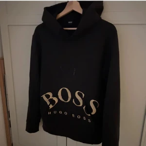 Svart hoodie från Hugo Boss - Snygg svart hoodie från Hugo Boss med stor logga framtill. Perfekt för en avslappnad stil. Den har långa ärmar och en klassisk huva. Passar bra till både jeans och träningsbyxor.