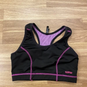 Sporttopp  - Snygg svart sporttopp från Kappahl med lila meshdetaljer och kontrastsömmar. Perfekt för träning med sin bekväma passform och andningsbara material. Toppen har en racerback-design för extra stöd. Storlek S/M