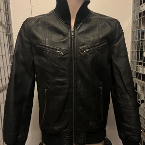 Svart skinnjacka från Poco Loco - Snygg svart skinnjacka från Poco Loco med dragkedja framtill och två bröstfickor med dragkedjor. Jackan har en klassisk look med ribbade muddar vid ärmslut och nederkant. Perfekt för en cool och stilren outfit.