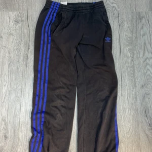 Svarta mjukisar från Adidas - Snygga svarta träningsbyxor från Adidas med klassiska blå ränder längs sidorna och Adidas-logga på benet. Byxorna har en elastisk midja för extra komfort. Perfekta för träning eller en avslappnad stil.