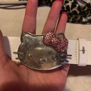 Vitt bälte med Hello Kitty-spänne - Snyggt vitt bälte med ett stort Hello Kitty-spänne i silver och rosa detaljer. Perfekt för att ge en lekfull touch till din outfit. Spännet har en glittrig rosett som verkligen sticker ut. har gjort egna hål i den, går enkelt att lägga till fler håll om man vill!