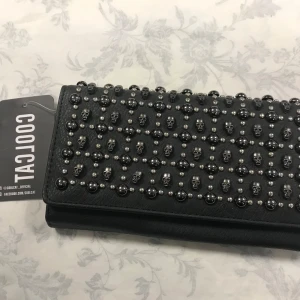 Svart plånbok med dödskallar  - Cool svart plånbok dekorerad med små dödskallar och nitar. Perfekt för att ge din stil en edgy touch. Plånboken har flera fack och en dragkedja inuti för säker förvaring av dina kort och kontanter.  Helt ny 