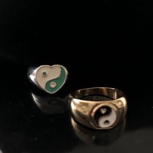 Två ringar med yin-yang-motiv - Säljer två ringar med yin-yang-design. En ring är i silver med ett hjärtformat motiv i vitt och grönt. Den andra ringen är i guld med ett runt motiv i svart och vitt. Perfekta för att ge en unik touch till din stil.