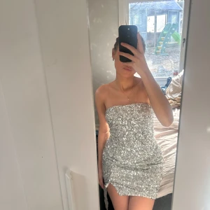 Glittrig axelbandslös klänning - Snygg och glittrig axelbandslös klänning i silver. Perfekt för en kväll ute med vänner eller en speciell tillställning. Klänningen har en tight passform som framhäver figuren.