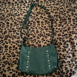 Mörkgrön axelväska med nitar - Snygg mörkgrön axelväska med coola nitar på framsidan. Väskan har en justerbar axelrem.