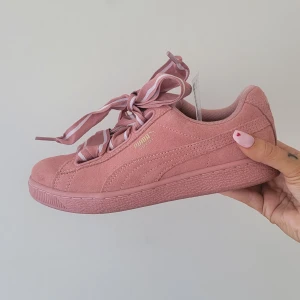 Rosa mocka sneakers från Puma - Säljer ett par snygga rosa sneakers från Puma i mocka. Skorna har breda snören med vita detaljer och en klassisk sula med mönster. Perfekta för en stilren look.