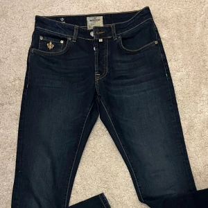 Mörkblå jeans från Morris - 29 32 Snygga mörkblå jeans från Morris med broderad detalj på fickan och klassisk femficksdesign. Jeansen har en rak passform och är tillverkade i ett slitstarkt denimtyg. Perfekta för en stilren look.