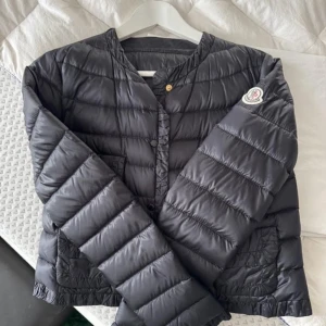 Svart dunjacka från Moncler - Säljer en stilren svart dunjacka från Moncler. Jackan har en quiltad design med knappar framtill och en rund halsringning. Perfekt för kyligare dagar och ger en lyxig känsla. Moncler-logga på ärmen ger en extra touch av exklusivitet. Storlek 36