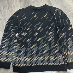 Boss tröja  - Snygg svart tröja från Boss med ett unikt geometriskt mönster i beige och grått. Tröjan har långa ärmar och en rund halsringning, perfekt för en stilren look.