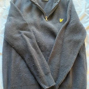 Mörkblå stickad tröja från Lyle & Scott - Säljer en stilren mörkblå stickad tröja från Lyle & Scott med dragkedja och broderad gul logotyp på bröstet. Perfekt för kyligare dagar med sin ribbade design och långa ärmar. Passar både till vardags och mer uppklädda tillfällen.