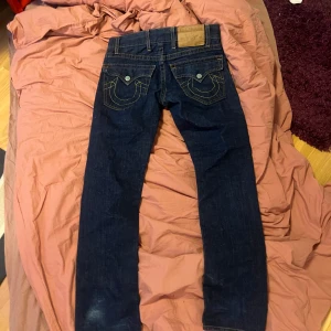 Mörkblå jeans med kontrastsömmar - Snygga mörkblå jeans från True Religion med ikoniska kontrastsömmar och detaljerade bakfickor. Jeansen har en klassisk femficksdesign och knappar med märkets logga. Perfekta för en stilren look.