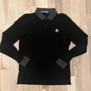 Svart långärmad pikétröja från Moncler - Säljer en stilren svart långärmad pikétröja från Moncler med grå krage och muddar. Tröjan har stl S