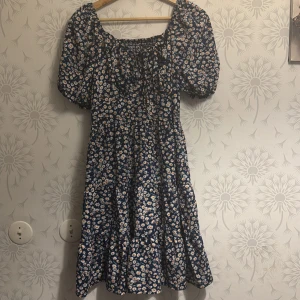 Blommig klänning med puffärmar - Charmig blommig klänning i mörkblått med vita och rosa blommor. Den har puffiga kortärmar och en smockad överdel som ger en fin passform. Perfekt för sommarens alla tillfällen. Liten i storlek som i storlek M/L