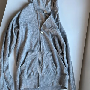 Grå munkjackan med stjärndetaljer - Säljer en grå munkjackan med dragkedja och huva. Den har en stjärna i paljetter på bröstet och texten 'I AM UNIQUE' med en stjärna på ryggen. Perfekt för en avslappnad stil. STORLEK 146/152