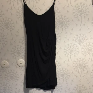 Svart klänning med smala axelband - Elegant svart klänning med smala axelband och v-ringad hals. Klänningen har en draperad detalj på sidan som ger en fin silhuett. Perfekt för en stilren look.