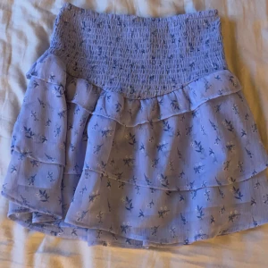 Blommig volangkjol - Söt blommig volangkjol i ljusblått med elastisk midja. Perfekt för en somrig look med sina flera lager av volanger och små blommönster. Kjolen är kort och ger en luftig känsla.