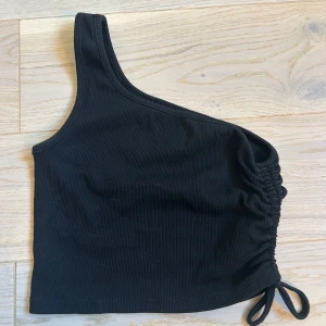 Svart oneshoulder topp - Snygg svart kroppad oneshoulder topp med ribbad textur och inspänningsbar knytning i sidan. Perfekt för en trendig look. Eftersom att lappen är bortklippt så vet jag inte storlek men skulle gissa på S