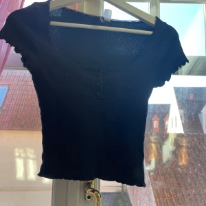 Svart ribbad topp från H&M Divided - Säljer en svart ribbad topp från H&M Divided i storlek XS. Toppen har korta ärmar och knappar framtill. Perfekt för en stilren look.