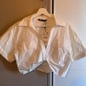 Vit croppad skjorta från Zara - Snygg vit croppad skjorta från Zara med korta ärmar och två bröstfickor. Perfekt för en trendig look. Skjortan har en klassisk krage och knappar framtill. Passar perfekt till både jeans och kjol för en stilren outfit. Liten i storleken sitter mer som en s 