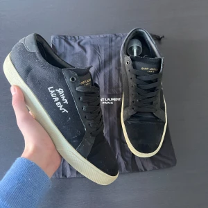 Saint Laurent skor - Snygga svarta sneakers från Saint Laurent med vit sula och klassisk snörning. Skorna har en stilren design med Saint Laurent-logga på sidan och tungan. Perfekta för en trendig och avslappnad look. Inga vidare defekter eller skador utan att på sulan där det ska stå saint Laurent har börjats skrapa bort som ni ser på bild 5, hör gärna av er vid frågor och funderingar