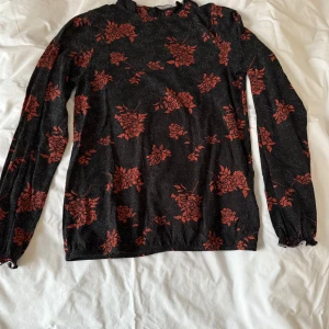 Svart blommig topp från House of Lola - Snygg svart topp med röda blommor från House of Lola. Tillverkad i mjukt Tencel-material med en liten andel elastan för stretch. Toppen har långa ärmar och en rund halsringning med en låg ståkrage i resår som gör att den sitter skönt. Toppen har en liten öppning bak som knäpps med knapp. Även i ärmarna finns en resår med diskret colang som en fin detalj. Toppen är sparsamt använd.
