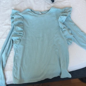 Ljusblå ribbad tröja från Serholt - Säljer en ljusblå ribbad tröja från Serholt med volangdetaljer längs ärmarna. Tröjan har långa ärmar och en rund halsringning. Perfekt för en stilren och bekväm look.
