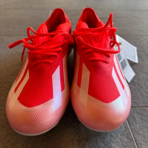 Röda fotbollsskor från Adidas - Snygga röda fotbollsskor från Adidas med vita detaljer och snörning. Skorna har en modern design med en lätt och flexibel sula för optimalt grepp på planen. Perfekta för den som vill ha stil och funktion i ett.