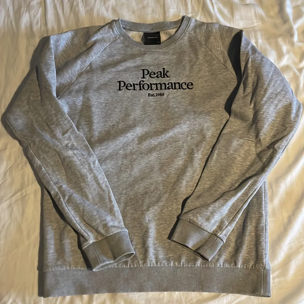 Säljer en grå sweatshirt från Peak Performance med svart text på bröstet. Tröjan har långa ärmar och ribbade muddar vid ärmslut och nederkant. Perfekt för en avslappnad stil. Priset kan sänkas vid en snabb affär. Storlek 170 typ XS.. Neuletakit & Villapaidat.