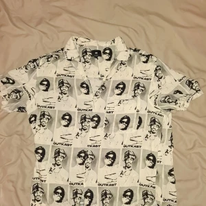 Vit kortärmad skjorta med Outkast-tryck från H&M - Säljer en cool vit skjorta från H&M med ett unikt svartvitt Outkast-tryck. Skjortan har korta ärmar och en avslappnad passform, perfekt för en avslappnad stil. Den är gjord i ett lätt och bekvämt material. Storleken är lite mindre än vad det står. Pris kan diskuteras.