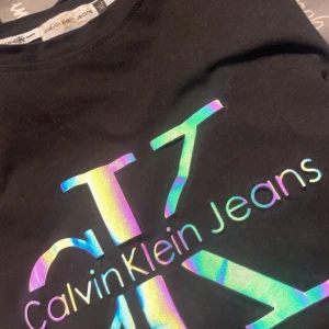 Svart t-shirt från Calvin Klein Jeans - Snygg svart t-shirt från Calvin Klein Jeans med ett coolt holografiskt tryck av logotypen på framsidan. Perfekt för en stilren och modern look. I storlek Xl