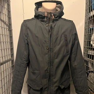 Grå parkajacka med teddyfoder - Säljer en grå parkajacka med mysigt teddyfoder. Jackan har en dragkedja och knappar framtill, samt flera praktiska fickor. Perfekt för kyliga dagar med sin långa passform och huva.