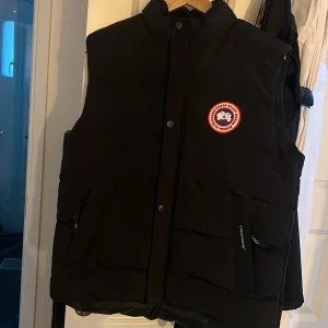 Svart dunväst från Canada Goose - Säljer en svart dunväst från Canada Goose med deras ikoniska logga på bröstet. Västen har en hög krage och stängs med dragkedja och knappar. Perfekt för kyliga dagar när du vill hålla värmen utan att kompromissa med stilen.