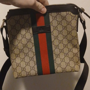 Gg väska kom privat för pris - Säljer en snygg beige axelväska från med klassiskt monogrammönster och röd-gröna ränder. Väskan har en justerbar axelrem och dragkedja upptill. Perfekt för att bära dina nödvändigheter med stil.