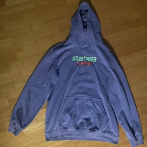 Ljuslila hoodie från H&M med tryck - Säljer en lila hoodie från H&M med ett färgglatt tryck på baksidan och texten 'Greetings' på framsidan. Hoodien har en stor ficka framtill och en bekväm passform. Perfekt för en avslappnad stil. Storlek M