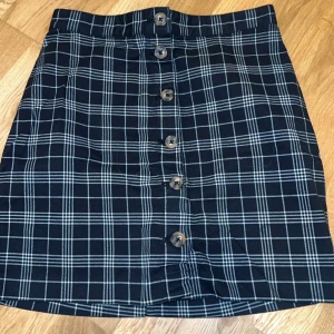 Rutig kjol från H&M Divided - Snygg rutig kjol från H&M Divided i svart och vitt. Kjolen har knappar framtill och en klassisk design som passar perfekt för olika tillfällen. Den är i storlek 36