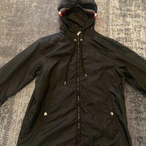  Moncler windbreaker  - Säljer en svart vindjacka från Moncler. Jackan har en justerbar huva med röd och vit randdetalj, dragkedja framtill och praktiska fickor med knappar. Perfekt för blåsiga dagar. Storlek M 