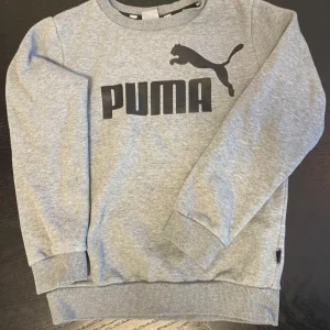 Grå sweatshirt från Puma - Säljer en stilren grå sweatshirt från Puma med deras ikoniska logga i svart på bröstet. Tröjan har långa ärmar och ribbade muddar vid ärmslut och nederkant. Perfekt för en avslappnad look.