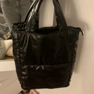 Svart quiltad axelväska - Snygg svart quiltad axelväska i syntetmaterial. Perfekt för att bära med sig det nödvändigaste med stil. Väskan har praktiska handtag och en dragkedja för säker förvaring.