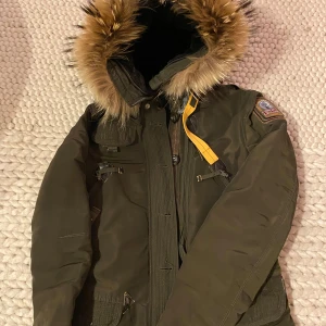 Grön parkajacka från Parajumpers - Säljer en grön parkajacka från Parajumpers med pälsfodrad huva och flera praktiska fickor. Jackan har en broderad patch på ärmen och dragkedja framtill. Perfekt för kalla vinterdagar        