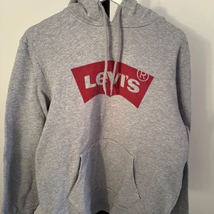 Grå hoodie från Levi's - Säljer en klassisk grå hoodie från Levi's med det ikoniska röda logotyptrycket på framsidan. Tröjan har en bekväm känguruficka och justerbar huva med dragsko. Perfekt för en avslappnad stil.nypris 650kr
