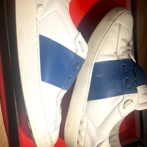 Valentino Garavani sneakers i vitt och blått - Snygga Valentino Garavani sneakers i vitt och blått med klassisk design. Skorna har en bred blå rem över mitten och subtila detaljer som ger en lyxig känsla. Dom är använda men forfarande i ett  bra skick.  Skorna passar bra för någon som har storlek 39 eftersom att passformen är rätt så bred och passar in sig efter ett tag i ens egna fot form. Fråga gärna frågor om du har några, och priset kan såklart Diskuteras. 