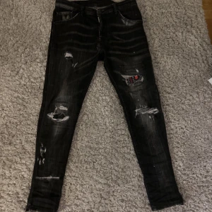 Svarta jeans med slitningar - Snygga svarta jeans med slitningar och en unik patchdetalj på ena benet. Perfekta för en trendig look med en tight passform.